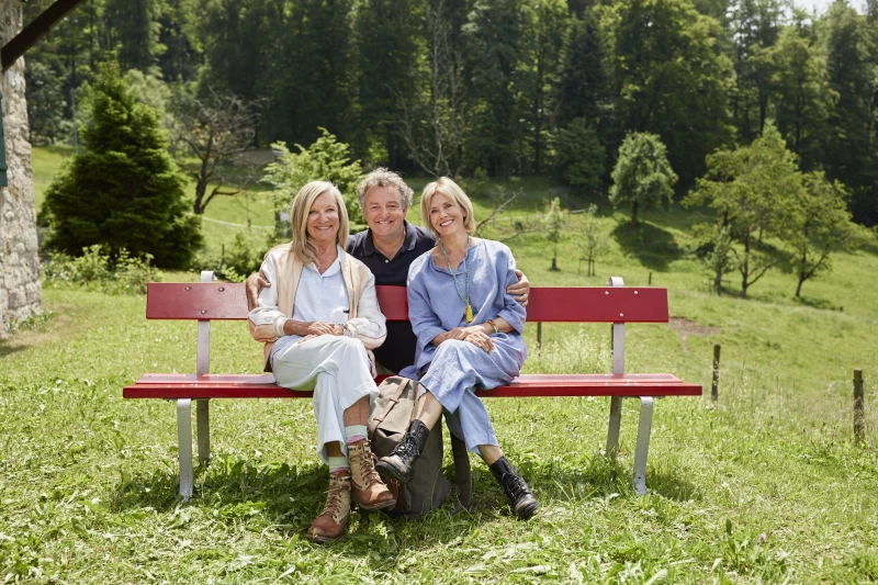 Drei Personen sitzen auf einer roten Bank in der Natur. 
